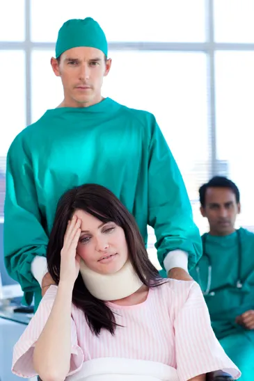 A doctor in green scrubs and cap pushes a female patient in a wheelchair who is wearing a neck brace and holding her forehead.