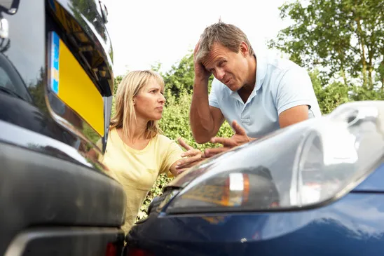 Two individuals are standing between the damaged fronts of two cars following a traffic accident.