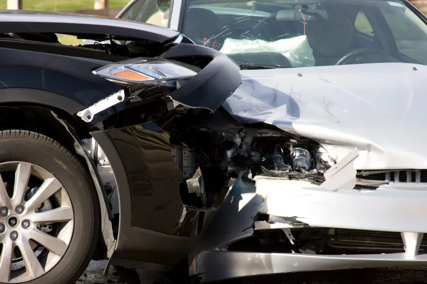 A head-on car collision between a dark vehicle on the left and a white vehicle on the right with a severe front-end damage and deployed airbags.