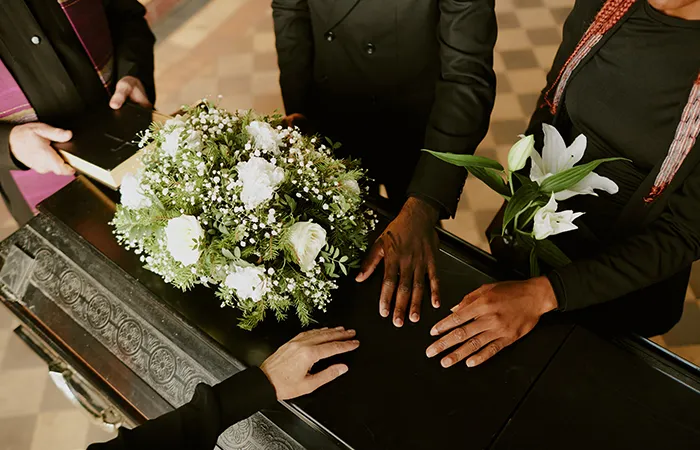 A black casket covered with a white floral arrangement, with several hands resting on the surface during a funeral service.
