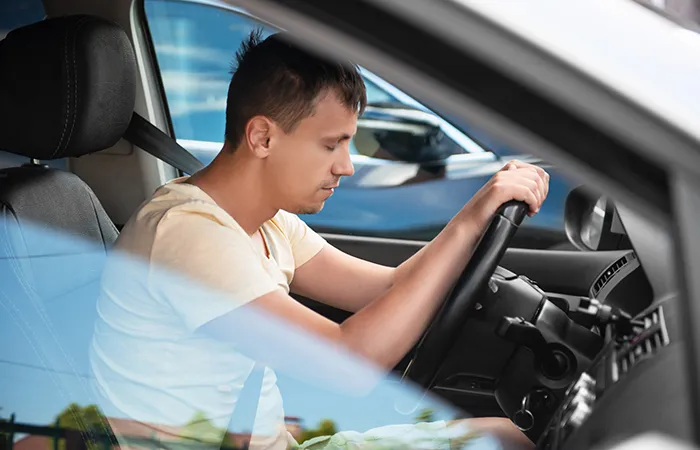 A young man with his eyes closed is sitting in the driver's seat of a car with both hands on the steering wheel, appearing sleepy or drowsy.