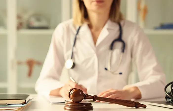 A wooden gavel resting on a sound block in the foreground, with a woman in a white doctor's coat and stethoscope writing at a desk.