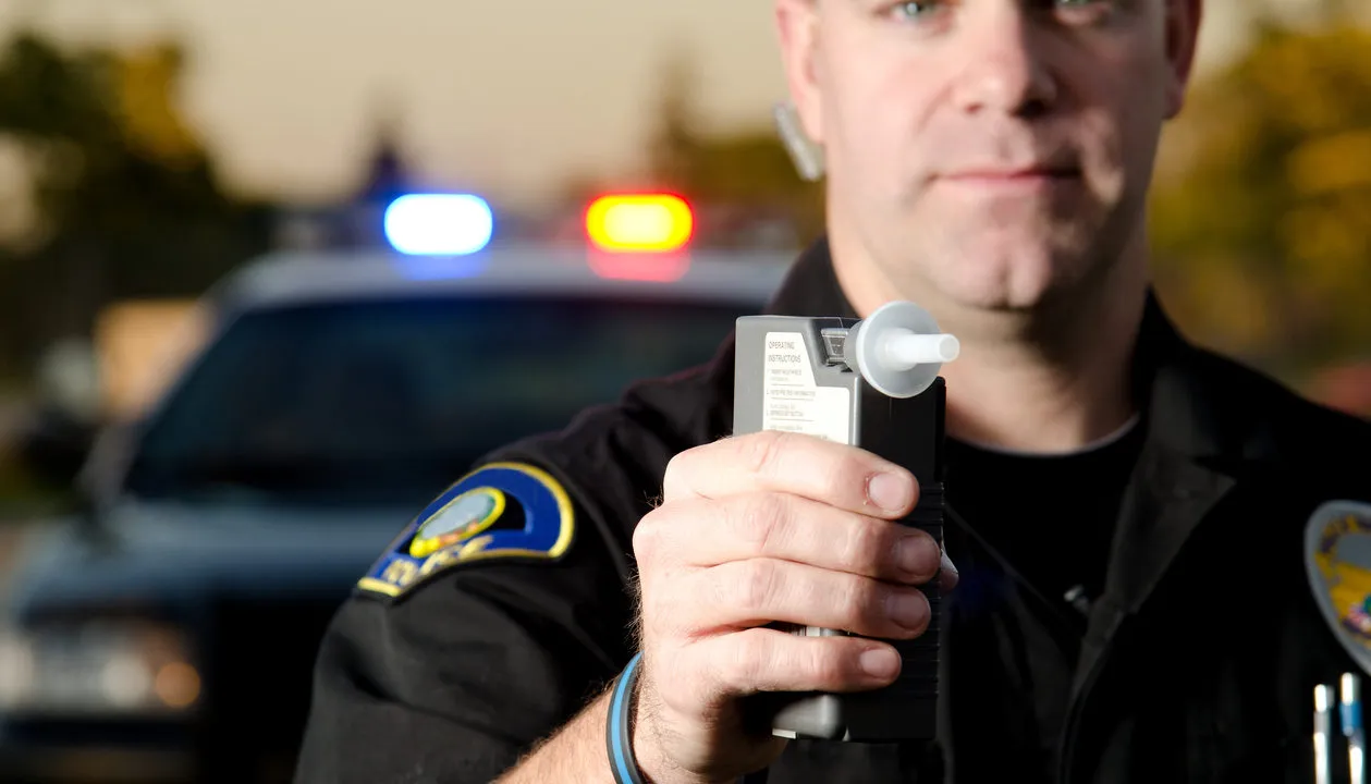 A police officer in a black uniform is holding a portable breath test device toward the camera.