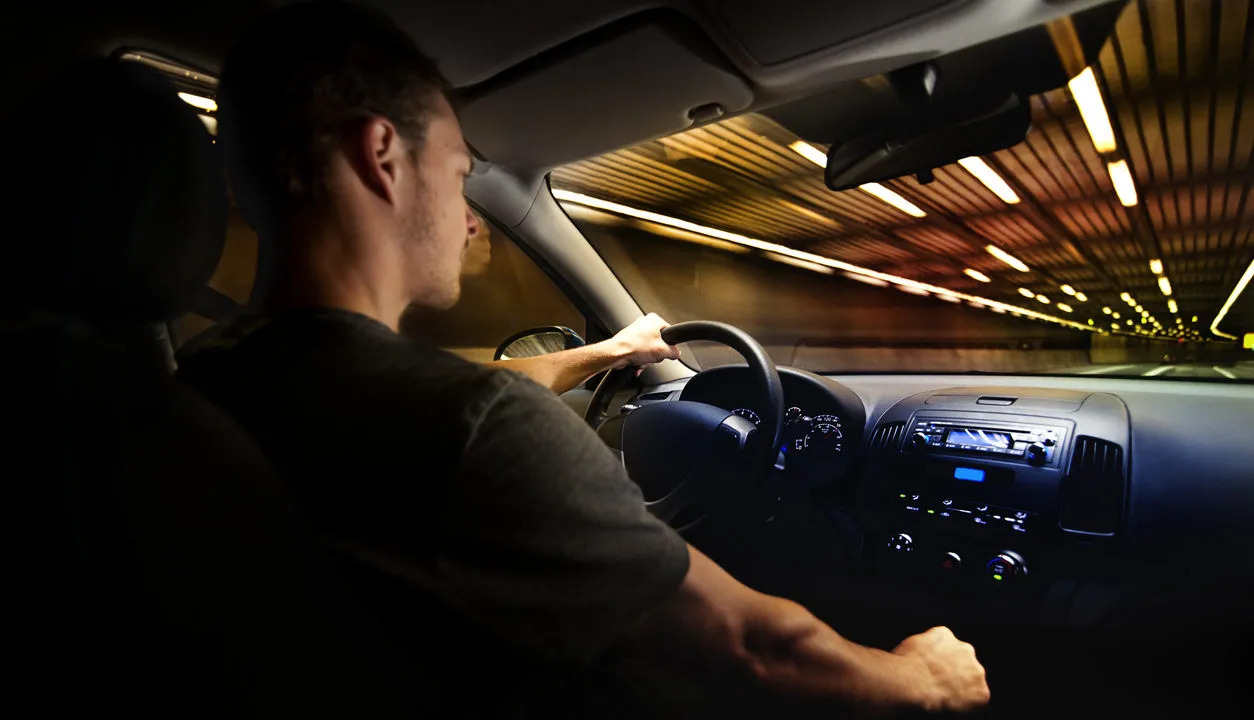 A man driving a car through a brightly lit tunnel at night, viewed from the passenger seat.