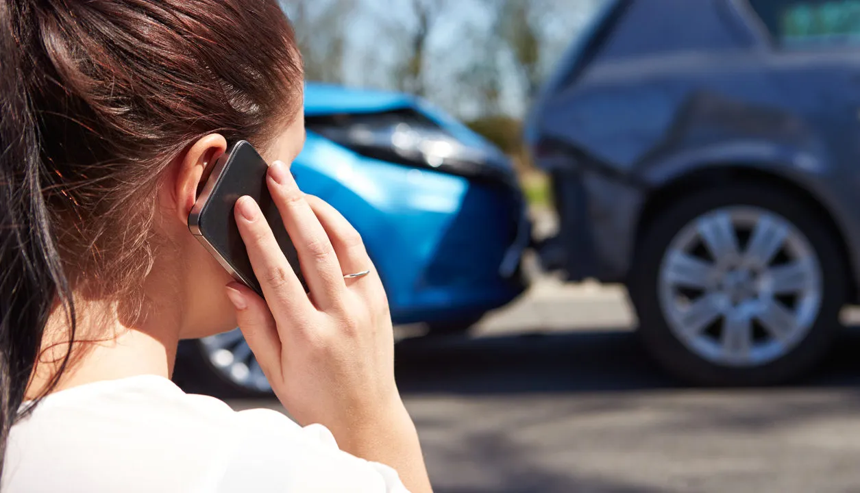 A woman talking on a cell phone in the front of the damaged car in rear end accident.