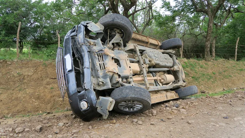The overturned car on a rough, dirt, and gravel roadside, next to a grassy ditch and a wooden fence.