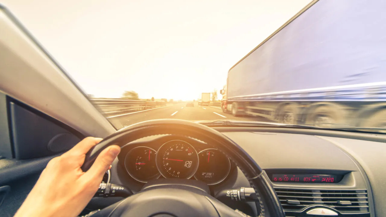 A hand is visible on the steering wheel, and the dashboard, with a fast-moving semi-truck is passing on the right.