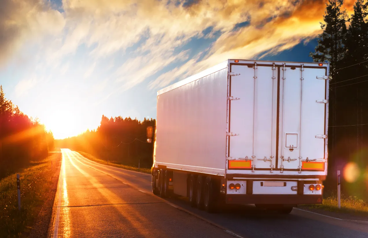 A white semi-trailer truck drives down a sun-drenched road, heading towards the bright sunset.