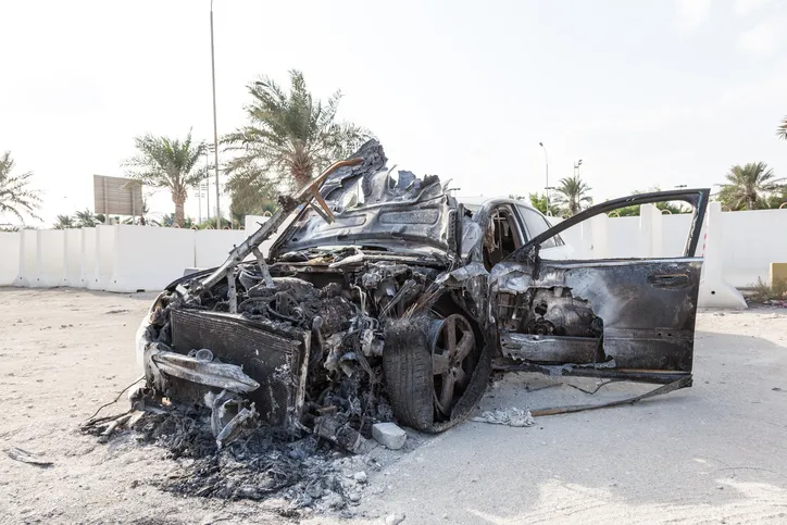 A black, charred, and heavily burnt sedan is resting on a dirt shoulder next to a white concrete barrier, likely after a fire or explosion.