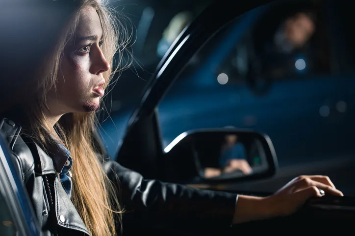 a young woman with long hair and a black leather jacket sitting in the driver's seat of a car, with bruises and cuts on her face.