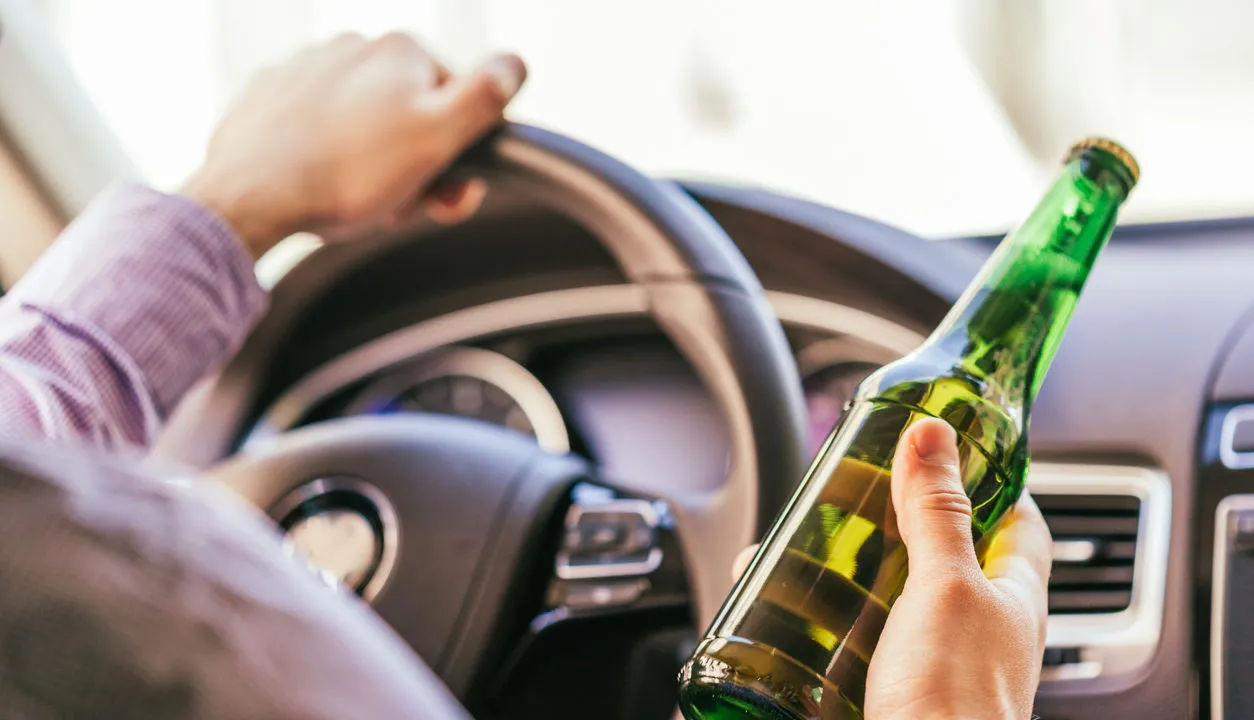 A person's hands holding the steering wheel of a car and the other hand holding an unopened green glass beer bottle.