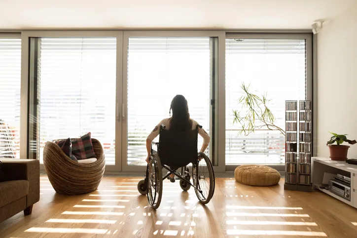 A woman with long dark hair sitting in a wheelchair and facing a large glass sliding door.