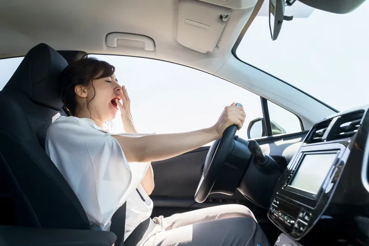 A woman is yawning widely and stretching her arm out while driving a car on a sunny day.