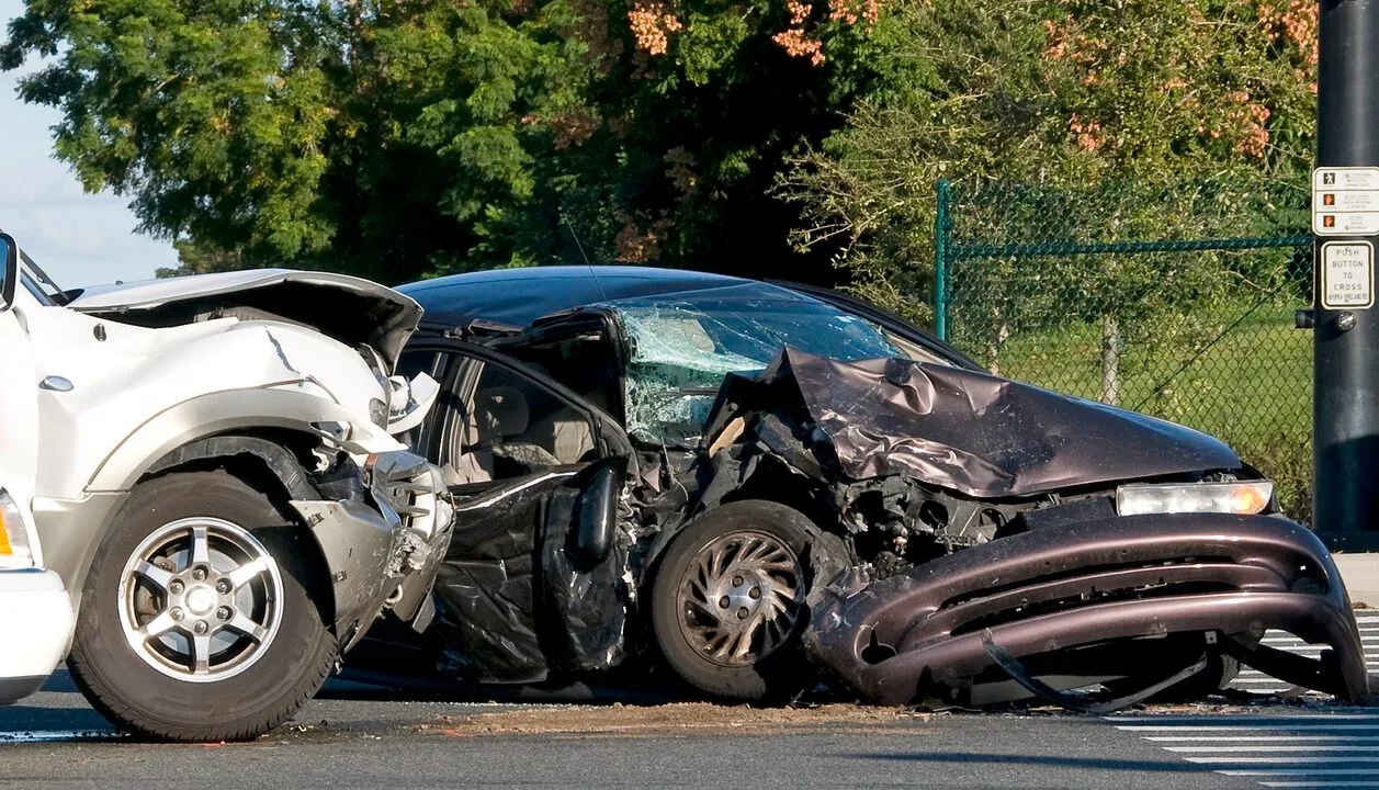 A head-on car accident between a partially visible white SUV on the left and a heavily damaged dark purple or black sedan on the right.