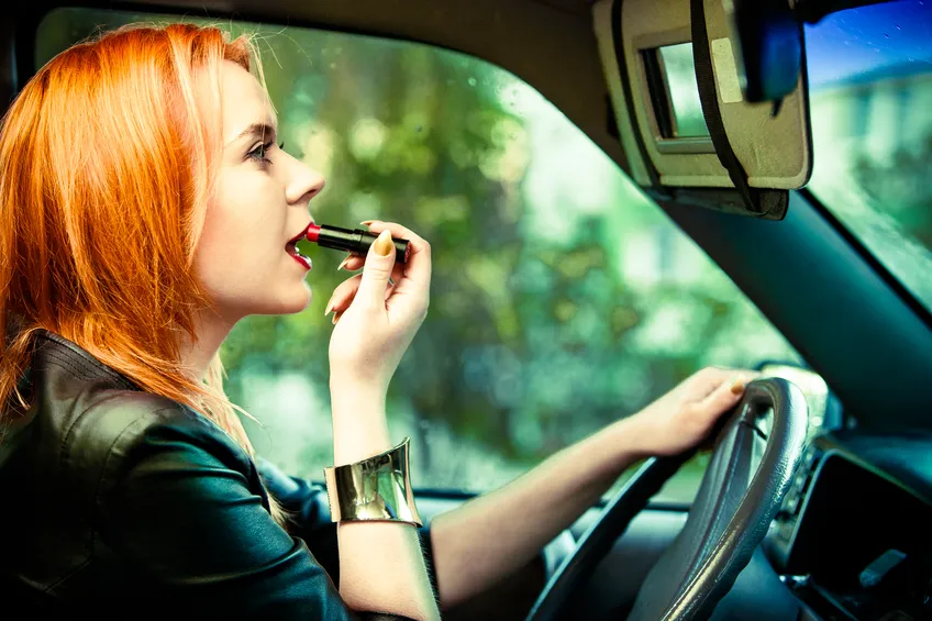 Woman with orange hair applies red lipstick while driving a vehicle.
