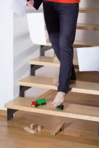 Barefoot man tripping on a toy train on the stairs while holding papers, suggesting a slip-and-fall accident.