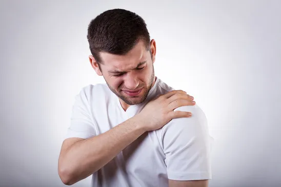A young man in a white t-shirt clutching his right shoulder with a painful and grimacing expression.