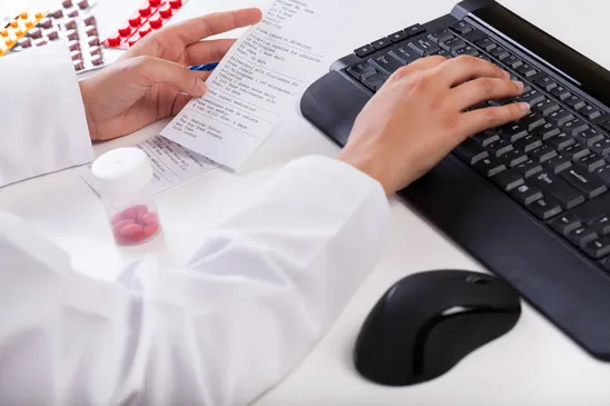 A pharmacist's hands working on a computer keyboard while reviewing a paper prescription with a bottle of red and white pills nearby.