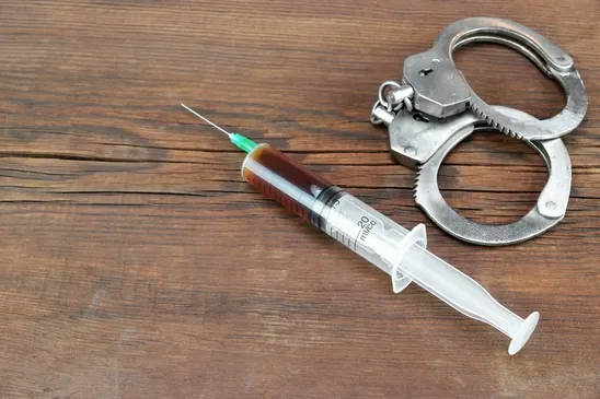 Syringe filled with brown liquid next to a pair of handcuffs on a wooden table.