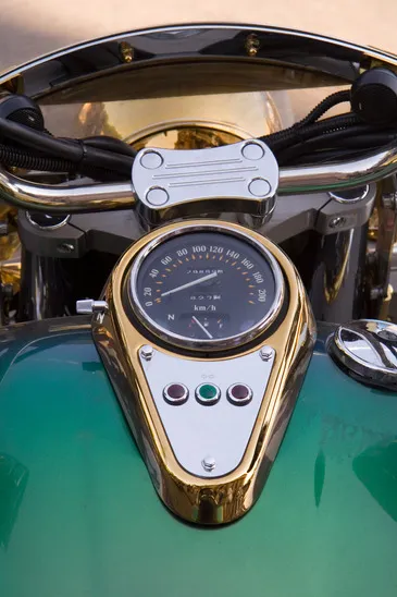 A motorcycle's dashboard, featuring a speedometer and warning lights and gold trim on a bright green fuel tank.