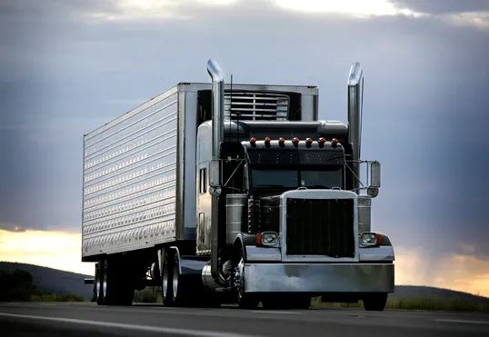 Black semi-truck with a silver refrigerated trailer driving on a highway under a dramatic cloudy sky at sunset.