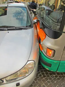 Severely damaged silver hatchback smashed against a bus/large vehicle on the road.
