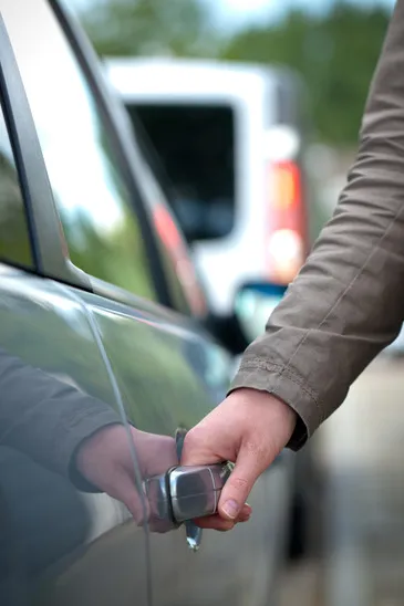 A person's hand in a tan jacket pulling the silver handle to open a dark car door.
