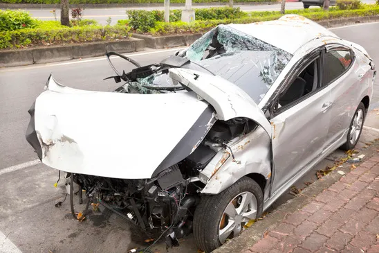 A severely damaged silver car pulled up against a curb and sidewalk following an accident.