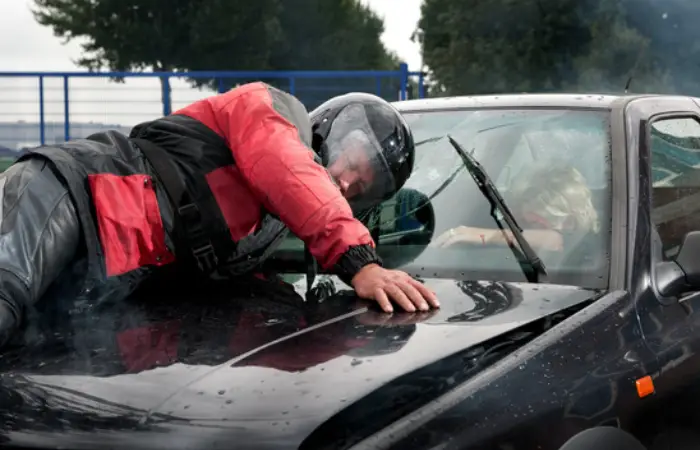 A man wearing a motorcycle helmet lies on the hood of a car after being crushed in a collision.