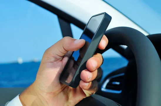 A person's hand holding a black smartphone while driving, with one hand on the steering wheel and the ocean visible through the car window.