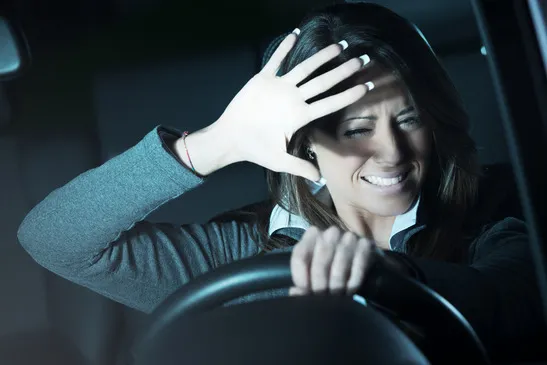 A young woman in the driver's seat of a car is holding the steering wheel and raising her right hand to shield her eyes from a bright light.