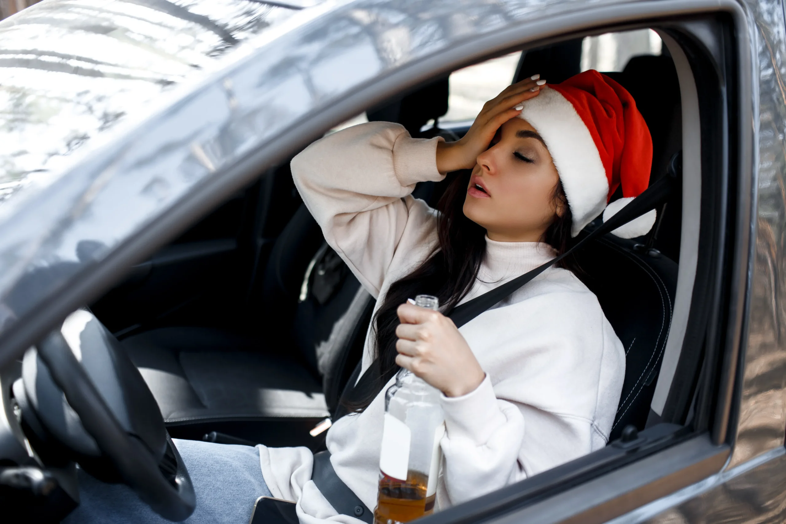 A young woman in a Santa hat, appearing unwell or intoxicated, is sitting in the driver's seat of a car, holding a bottle of alcohol and touching her forehead.