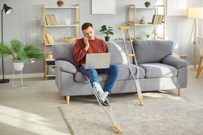 Injured young man with bandaged leg sitting on sofa, working on laptop and talking on mobile phone with crutches lying nearby.